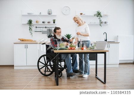 Senior husband in wheelchair and wife mixing chopped vegetables in bowl. Senior husband in wheelchair and wife mixing chopped vegetables in bowl. 110384335