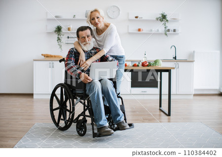 Charming old couple of man in wheelchair with laptop and wife hugging him. Charming old couple of man in wheelchair with laptop and wife hugging him. 110384402