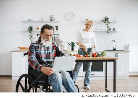 Old man sits in wheelchair and works on laptop while his wife prepares healthy salad. Old man sits in wheelchair and works on laptop while his wife prepares healthy salad. 110384406
