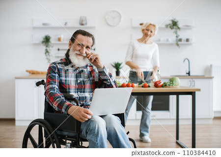 Old man sits in wheelchair and works on laptop while his wife prepares healthy salad. 110384410