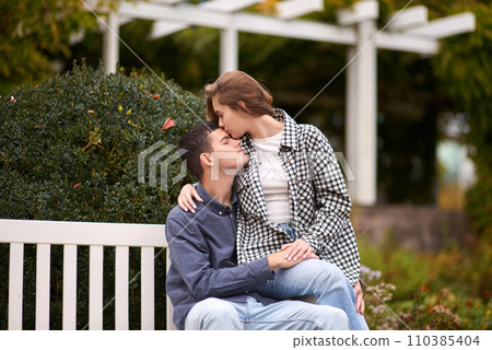 Autumn Romance: Young Couple Embracing and Kissing on Park Bench. Young couple kissing and rejoices at the lake. lovely young couple kissing outdoors in autumn. Embraced in Autumn's Warmth: Couple's 110385404