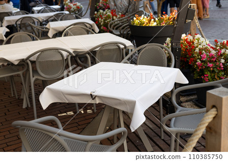 Dining outdoors. Outdoor street cafe tables ready for service. Empty cafe terrace with table and chair. Table of outdoor cafe on sidewalk with colorful plants and flowers 110385750