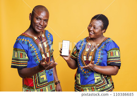 Smiling black couple wearing ethnic closes showing smartphone with white blank screen mockup. Cheerful man and woman presenting mobile phone with empty touchscreen for app advertising Smiling black couple wearing ethnic closes showing smartphone with white blank screen mockup. Cheerful man and woman presenting mobile phone with empty touchscreen for app advertising 110386685