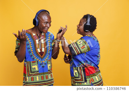 Cheerful black man and woman couple chatting while listening to music in wireless headphones. Mature boyfriend and girlfriend using mobile phone and talking while enjoying sound in earphones Cheerful black man and woman couple chatting while listening to music in wireless headphones. Mature boyfriend and girlfriend using mobile phone and talking while enjoying sound in earphones 110386746