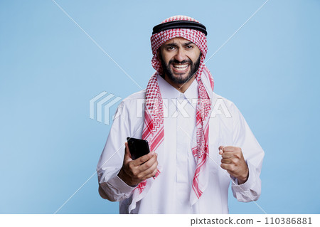 Happy arab reading good news on mobile phone and looking at camera with excited expression. Cheerful muslim man in thobe using smartphone and showing winner gesture with clenched fist portrait Happy arab reading good news on mobile phone and looking at camera with excited expression. Cheerful muslim man in thobe using smartphone and showing winner gesture with clenched fist portrait 110386881