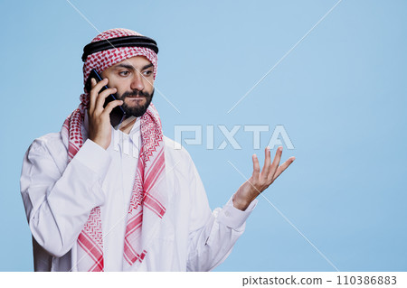 Arab man posing in traditional clothes while speaking on smartphone and making gesture with hand. Muslim person having communication on mobile phone while standing in studio Arab man posing in traditional clothes while speaking on smartphone and making gesture with hand. Muslim person having communication on mobile phone while standing in studio 110386883