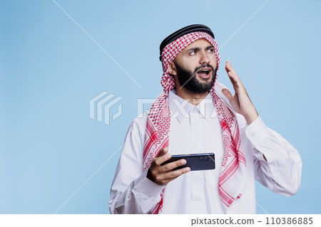 Upset muslim man wearing traditional islamic clothes loosing mobile game while enjoying online leisure in studio. Unhappy arab gamer in ghutra and thobe playing videogame on phone Upset muslim man wearing traditional islamic clothes loosing mobile game while enjoying online leisure in studio. Unhappy arab gamer in ghutra and thobe playing videogame on phone 110386885