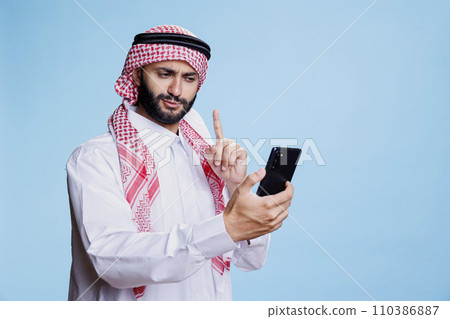 Muslim man dressed in traditional headscarf and thobe while speaking in videoconference using smartphone. Arab pointing with finger while chatting in online meeting on mobile phone Muslim man dressed in traditional headscarf and thobe while speaking in videoconference using smartphone. Arab pointing with finger while chatting in online meeting on mobile phone 110386887