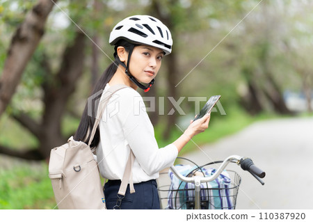 一位戴著自行車頭盔的年輕女子一邊拉著自行車一邊走，一邊看著智慧型手機上的地圖，檢查周圍的環境。 110387920