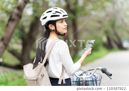 一位戴著自行車頭盔的年輕女子一邊拉著自行車一邊走,一邊看著智慧型手機上的地圖,檢查周圍的環境。 一位戴著自行車頭盔的年輕女子一邊拉著自行車一邊走,一邊看著智慧型手機上的地圖,檢查周圍的環境。 110387922