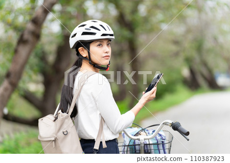 A young woman wearing a cycling helmet walks while pulling her bicycle while looking at a map on her smartphone and checking her surroundings. A young woman wearing a cycling helmet walks while pulling her bicycle while looking at a map on her smartphone and checking her surroundings. 110387923