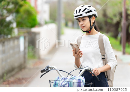 一位戴著自行車頭盔的年輕女子一邊拉著自行車一邊走,一邊看著智慧型手機上的地圖,檢查周圍的環境。 一位戴著自行車頭盔的年輕女子一邊拉著自行車一邊走,一邊看著智慧型手機上的地圖,檢查周圍的環境。 110387925