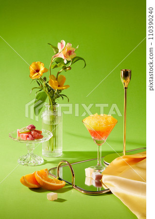 Front view of delicious smoothie cups, orange slices and jams on mirror sheet decorated with glass vase of flower and yellow silk fabric on a green background. Creative scene for advertising Front view of delicious smoothie cups, orange slices and jams on mirror sheet decorated with glass vase of flower and yellow silk fabric on a green background. Creative scene for advertising 110390240