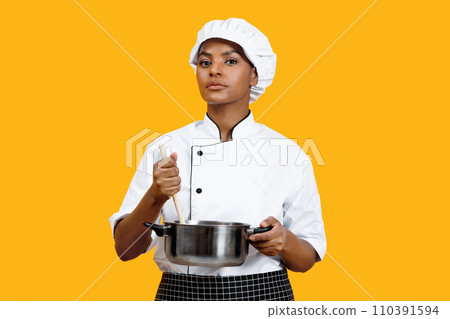 Serious black female chef holding wooden spoon and stainless steel pot 110391594