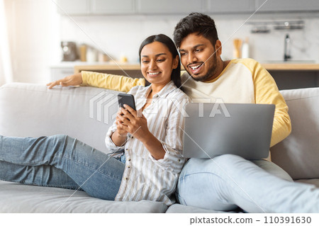 Engaged indian couple relaxing on couch with smartphone and laptop Engaged indian couple relaxing on couch with smartphone and laptop 110391630