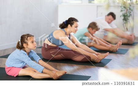 Preschool child performs back stretching pose, Paschimottasana. Family does hatha yoga in background 110391631