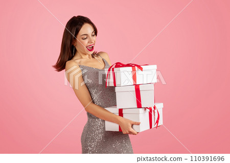 Joyful young woman in a shiny sequin dress excitedly holding stacked white gift boxes with red ribbons Joyful young woman in a shiny sequin dress excitedly holding stacked white gift boxes with red ribbons 110391696