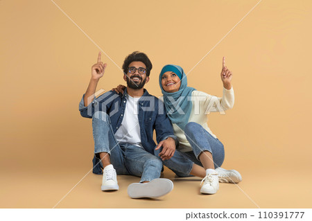 Joyful muslim couple sitting together against beige background and pointing upwards Joyful muslim couple sitting together against beige background and pointing upwards 110391777