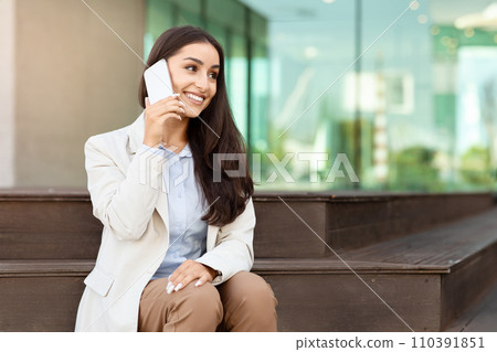 Smiling businesswoman in a light blazer talking on a smartphone while sitting on an outdoor bench 110391851