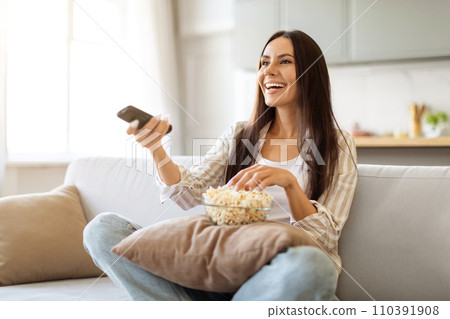 Woman enjoying movie night at home, holding a remote and eating popcorn 110391908