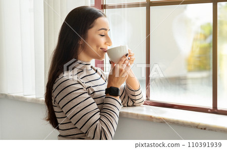 Contented young woman with sleek hair in a smartwatch and striped shirt savors her coffee 110391939