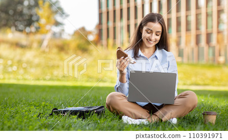Smiling young professional woman multitasking with a laptop on her lap, enjoying a sandwich Smiling young professional woman multitasking with a laptop on her lap, enjoying a sandwich 110391971