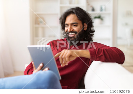 Cheerful young indian man sitting on couch, using digital tablet 110392076
