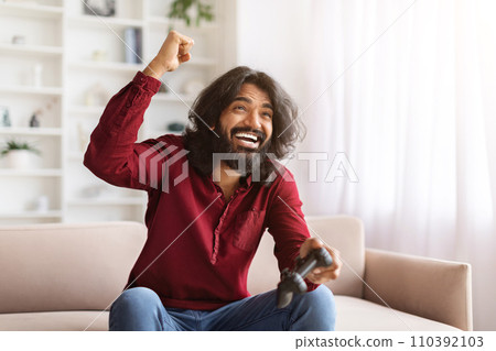 Excited emotional indian man gamer sitting on couch at home Excited emotional indian man gamer sitting on couch at home 110392103