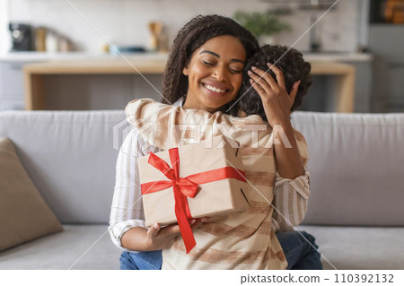 Joyful black mother holding wrapped gift box and affectionately embracing her son Joyful black mother holding wrapped gift box and affectionately embracing her son 110392132