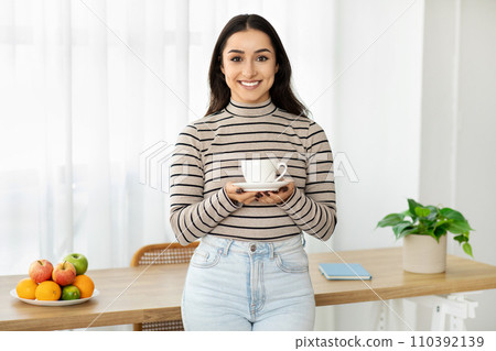 Smiling young woman presenting a cup of coffee with both hands, standing in a kitchen 110392139