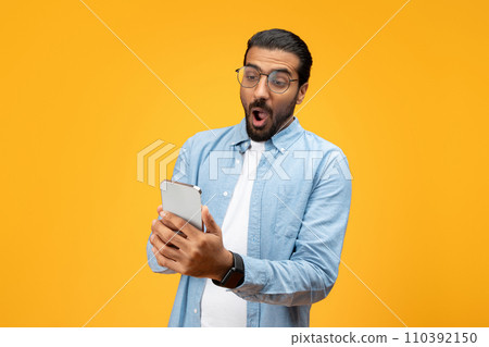 A startled man in a blue shirt stares at his smartphone with wide eyes and open mouth 110392150