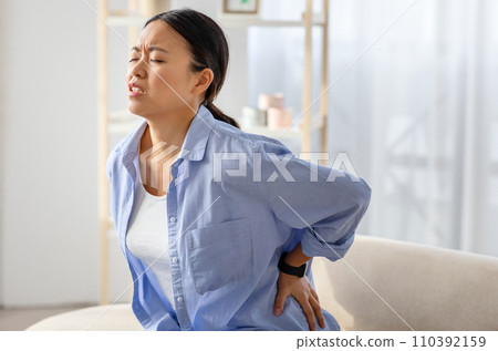 Chinese woman rubbing lower back, sitting on couch at home Chinese woman rubbing lower back, sitting on couch at home 110392159