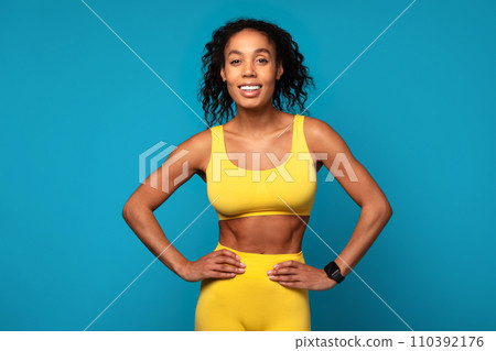 athletic black lady posing with hands on hips, blue backdrop 110392176
