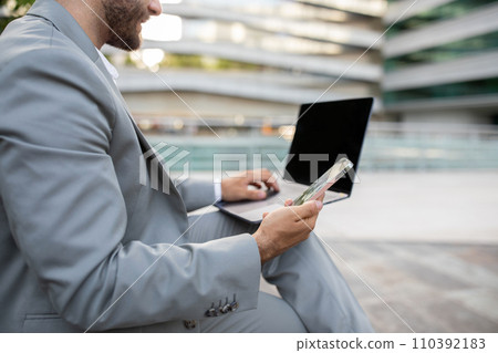 Unrecognizable young businessman using his smartphone while working on blank laptop outdoors Unrecognizable young businessman using his smartphone while working on blank laptop outdoors 110392183