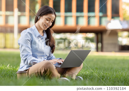 Content young woman in a blue shirt and brown pants enjoys working on her laptop while sitting cross-legged 110392184