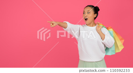 Shocked young asian woman with bright shopping bags pointing aside 110392233