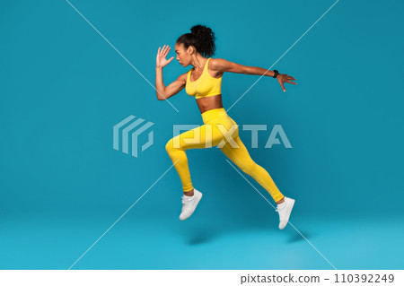 Athletic black woman leaping during dynamic workout on blue backdrop 110392249