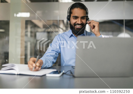 Cheerful indian man with headset working online on laptop 110392250