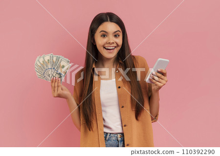 Cheerful teenage girl holding cash in one hand and smartphone in other Cheerful teenage girl holding cash in one hand and smartphone in other 110392290