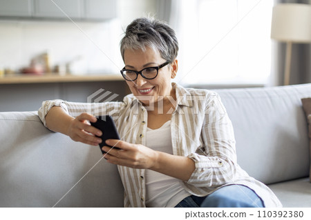 Smiling Senior Woman Using Smartphone While Relaxing On Couch At Home 110392380