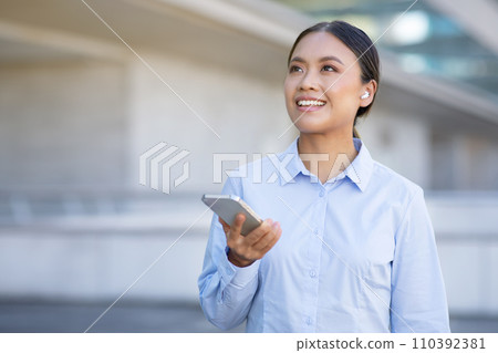 Korean business lady wearing earbuds holds smartphone looking aside outdoor 110392381