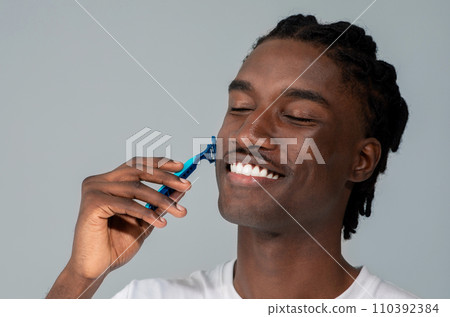 Cheerful handsome black man shaving his face with blue razor Cheerful handsome black man shaving his face with blue razor 110392384