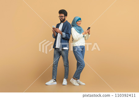 Modern arab muslim couple with their smartphones standing over beige background Modern arab muslim couple with their smartphones standing over beige background 110392411