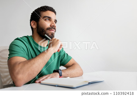 Pensive young man with a beard in a green shirt holding a pen to his chin Pensive young man with a beard in a green shirt holding a pen to his chin 110392418