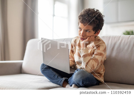 Pensive little black boy sitting with laptop on couch and looking away Pensive little black boy sitting with laptop on couch and looking away 110392459