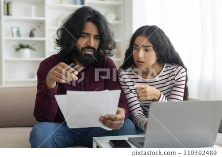Overwhelmed couple sitting together on couch, using laptop, holding papers Overwhelmed couple sitting together on couch, using laptop, holding papers 110392489