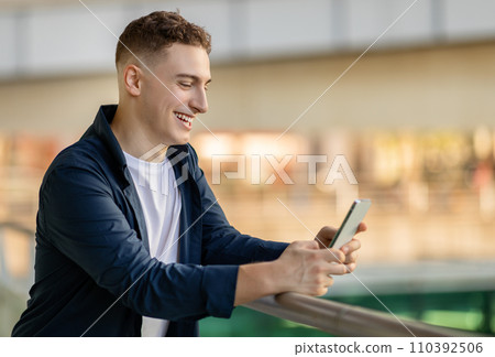 A smiling young man using a smartphone, leaning on a railing, dressed A smiling young man using a smartphone, leaning on a railing, dressed 110392506