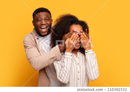Black man covering woman's eyes for surprise on yellow background 110392532