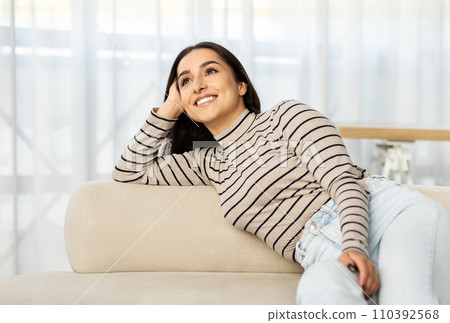 Pensive glad young arab woman lies on sofa in light living room Pensive glad young arab woman lies on sofa in light living room 110392568
