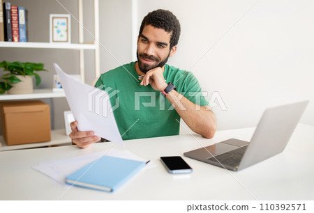 Contemplative man in a casual green t-shirt holds a pen to his chin while reviewing a document Contemplative man in a casual green t-shirt holds a pen to his chin while reviewing a document 110392571